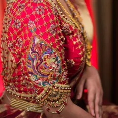 A close-up of the sleeve of our maroon bridal blouse, highlighting the complex zardosi and threadwork. The peacock motif is brought to life with a vibrant palette, set against a delicate net-like pattern of golden embroidery.