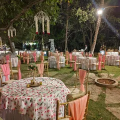An enchanting outdoor reception setup amidst the lush greenery of the ashram. The floral tablecloths, elegant chair drapes, and hanging chandeliers create a magical atmosphere for an evening celebration.