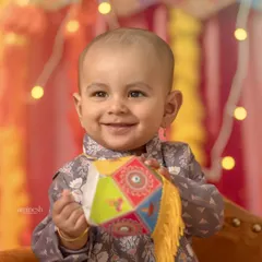 A close-up portrait of a smiling baby boy in traditional attire for a Diwali themed shoot. The warm, festive lights in the background add a celebratory glow.