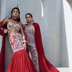 For the bride and her tribe, I offer coordinated yet distinct ensembles. This duo in crimson features a sculpted fish-cut lehenga and a pearl-embellished corset with tailored pants, both finished with dramatic crushed capes.