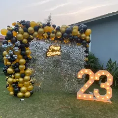 Celebrating a milestone birthday outdoors? This setup for a 23rd birthday features a shimmering silver sequin wall, a black and gold organic balloon garland, a custom neon sign, and large light-up marquee numbers.