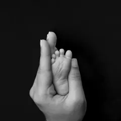 A minimalist black and white photo of a parent's hand holding their newborn's foot. The composition creates a heart-like shape, symbolizing love and protection.