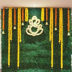A creative blend of traditional and modern, this backdrop features a green grass wall with hanging marigold garlands and a central Ganpati motif made from white jasmine flowers. The hanging bells add a divine touch.