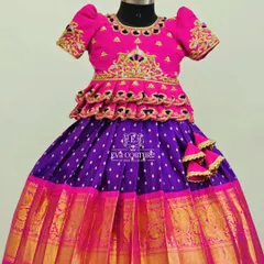 A vibrant pink and purple Pattu Pavadai. The blouse has a tiered peplum design and is detailed with rich gold embroidery, matching the wide zari border on the skirt.