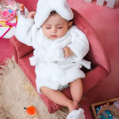 A fun and playful "spa day" theme. The baby is dressed in a tiny white bathrobe and slippers, lounging in a miniature chair for a truly unique and humorous newborn portrait.