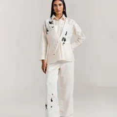 An off-white co-ord set featuring a structured, draped top and matching trousers, both adorned with an artistic black paint-splatter effect.