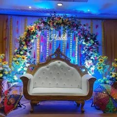 A colorful Haldi stage with a floral arch and a royal white sofa. The backdrop features a "Haldi" neon sign, and the setup is completed with decorative Rajasthani umbrellas.
