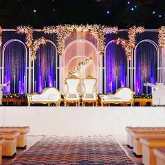 A sophisticated stage for a Sangeet or reception. The design features royal-style white and gold thrones, set against a modern arched backdrop with elegant floral arrangements and dramatic uplighting.