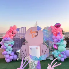 An outdoor mermaid-themed first birthday party set against a beautiful sunset. The decor features a large seashell-shaped backdrop, iridescent panels, and balloon garlands in shades of pink, purple, and teal, perfect for a magical under the sea adventure.