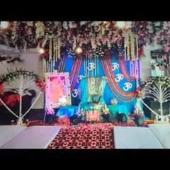 This Guruji Satsang stage decoration was designed with a vibrant blue color theme as requested by the client. The setup includes a floral canopy, a draped backdrop with marigold 'Om' symbols, and a decorated Aasan.