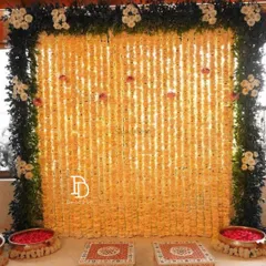 This elegant Mehndi backdrop features a full wall of light yellow flower strings, framed by a border of dense green foliage. The low decorated stools, or chowkis, provide traditional seating for the ceremony.
