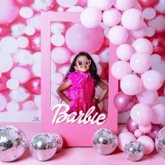 A vibrant 'Barbie' themed photoshoot for a stylish toddler. She poses confidently in a pink frame against a wall of pink balloons and disco balls, complete with a feather boa and sunglasses.