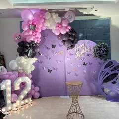 A beautiful butterfly theme for a 12th birthday. This setup includes a purple backdrop with butterfly decals, a balloon arch, and a large marquee number.