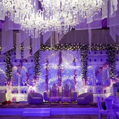 A breathtaking stage for an engagement ceremony, bathed in lavender light. The backdrop features intricate jaali work, floral adornments, and a custom neon hashtag, all under a canopy of crystal chandeliers.