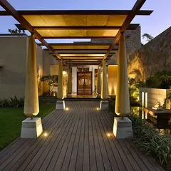 The entrance to C Farm in Chattarpur features a wooden deck walkway flanked by sculptural columns, leading to a grand door. The design uses a covered pergola to create a dramatic sense of arrival and transition from the outside world.
