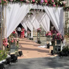 An elegant outdoor entrance featuring draped white fabric, floral arches, and crystal chandeliers, creating a soft and romantic pathway for a daytime wedding ceremony.