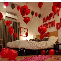 A cozy and romantic birthday room setup. The decor features red and black balloons, heart-shaped foils, and rose petals scattered on the floor and bed.