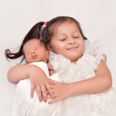 An angelic portrait of an older sister with her eyes closed in a happy smile as she lovingly hugs her newborn sibling, capturing a moment of pure sibling love.