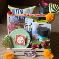 A fun and colorful Raksha Bandhan hamper in a wooden crate. It includes popcorn, a water bottle, sweets, and other treats, with cute stickers for a brother and sister-in-law.