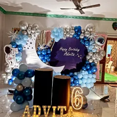 An out-of-this-world space-themed birthday decoration. This setup features a deep blue and silver balloon arch, a crescent moon cutout, and astronaut props to create a galactic adventure for the birthday star.