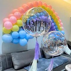 A pastel rainbow balloon arch over the sofa creates a dreamy backdrop for a birthday celebration, complemented by personalized confetti balloons.