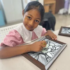 A young girl smiles as she works on her glass painting project, carefully filling in the design with vibrant colors.
