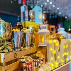 A close-up of the Gatsby bar's glowing details. The lit-up 'BAR' sign reflects on the mirrored surface, surrounded by ornate gold and copper glassware filled with aromatic garnishes like dried orange slices.