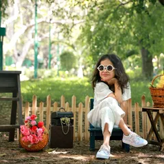 A stylish little girl posing confidently in a park. The natural setting with a rustic fence and baskets of flowers creates a charming scene for this outdoor fashion shoot.