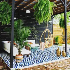 This covered patio design is all about boho-chic comfort. The space is defined by a bold patterned floor, a hanging swing chair, a plush daybed, and lots of hanging ferns, creating a relaxed and stylish outdoor living room.