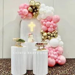 A beautiful baptism decoration for a baby girl, featuring a pink, white, and gold balloon arch. The setup includes a custom arched backdrop with a neon cross sign and fluted pedestals for desserts.