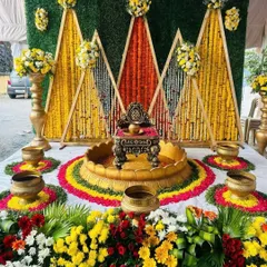 A colorful Haldi seating area with a backdrop of marigold strings arranged in geometric patterns against a green wall, surrounded by fresh flowers.