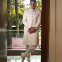 A sharp and stylish full-length portrait of a groom in a classic white sherwani. The natural light from the window and the confident pose create a sophisticated and timeless look for his wedding day.