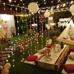 A full view of an elaborate backyard birthday party. The space is filled with string lights and paper lanterns, illuminating a long boho picnic table and a cozy teepee tent in the background.