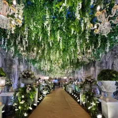 A Roman-inspired grand passageway with a ceiling of hanging greenery and white wisteria, lit by dozens of chandeliers. The aisle is lined with large floral urns, creating a path of epic proportions.