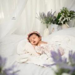 A newborn rests peacefully in a bed of soft white linens, framed by delicate lavender flowers. This lifestyle newborn photography approach creates a natural and timeless portrait.