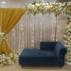 This engagement stage combines a white and gold floral arch with shimmering gold drapes and fairy lights. The setup includes floor candles and a royal blue sofa for a touch of color.