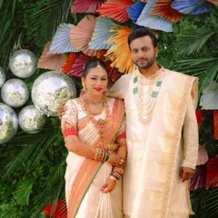 A full portrait of the wedding couple, showcasing their coordinated cream and red outfits against a festive backdrop. The details, from her bangles to his layered necklace, complete the picture-perfect look.