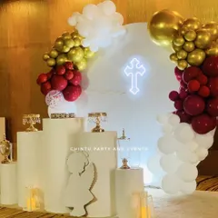 A First Holy Communion decoration in a striking red, gold, and white color scheme. The setup features a circular backdrop with a neon cross, an angel cutout prop, and pedestals for cake and other items.