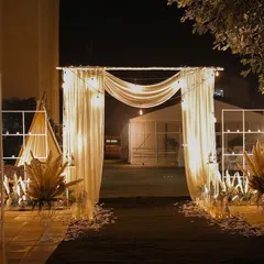 An ethereal entrance for a bohemian-themed night event. The path is lined with scattered petals and features a simple draped arch with warm Edison bulbs.