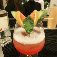 A beautifully garnished pink gin cocktail, ready to be served. The vibrant color, frothy top, and fresh garnishes of orange and basil showcase our attention to presentation and flavor.