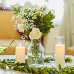Simple elegance for an at home celebration. A clear glass vase holds a delicate arrangement of white roses and baby's breath, flanked by glowing candles, creating a serene and beautiful centerpiece.