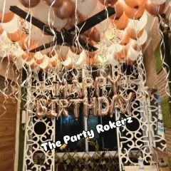 A surprise birthday setup in a room with rose gold and white balloons on the ceiling and "Happy Birthday" foil letters on the wall.