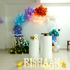This beautiful balloon ring decoration for an 8th birthday uses a rainbow of colors, from deep blues and purples to orange and white. The setup is completed with a neon sign, cake plinths, and illuminated marquee letters for the name 'RISHAAN'.