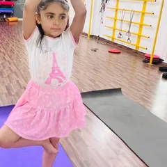 A young girl practices Vrikshasana (Tree Pose) on her purple yoga mat. This pose is fundamental for developing balance and focus, and she is doing it with great concentration.