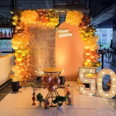 A grand 50th birthday celebration with a large "50" marquee number, a sequin and peach-toned backdrop, and a stunning arch of orange and gold balloons.