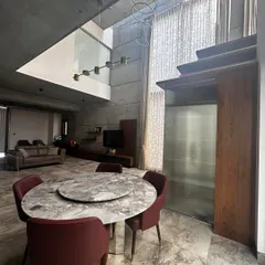 This open-plan dining and living area in a concrete-finished home showcases a double-height ceiling and expansive windows, creating a grand and airy atmosphere.