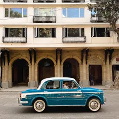 A blue and white Fiat, another classic, driving past the arched windows of a heritage building. This series celebrates the timeless appeal of vintage cars in Mumbai.