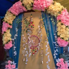 This arched backdrop combines yellow and pink artificial flowers with a two-tone fabric background. The hanging floral strings, bells, and a central decorative wheel create a layered and festive look suitable for any pooja.