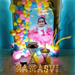 A colorful and personalized first birthday decoration. This setup includes a custom backdrop with the baby's photo, a vibrant balloon arch, and light-up marquee letters for the name.