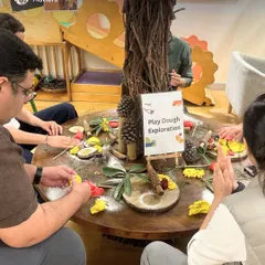 At our Parent Orientation, adults gather around a table to explore playdough and loose parts like pinecones and wood cuttings. This activity demonstrates how simple, natural materials can spark complex, imaginative play.